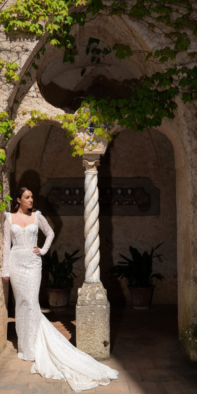 A bride in an elegant white gown standing near a twisted column under a vine-covered archway at Villa Cimbrone.