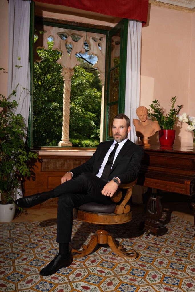 A groom in a black suit sitting in an elegant room with ornate tiled floors and a large window at Villa Cimbrone.