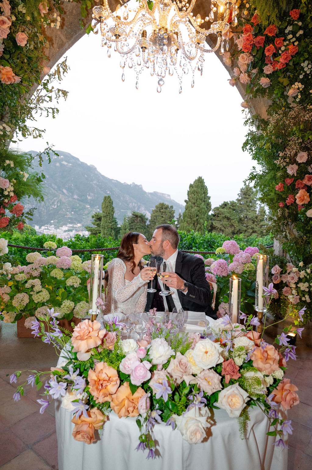 A bride and groom sharing a kiss at a floral-decorated table under a chandelier in the gardens of Villa Cimbrone.