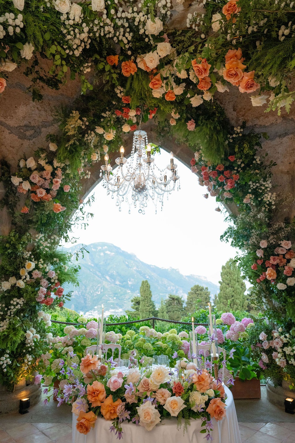 A beautifully decorated wedding table under a floral arch and chandelier overlooking the Amalfi Coast at Villa Cimbrone.