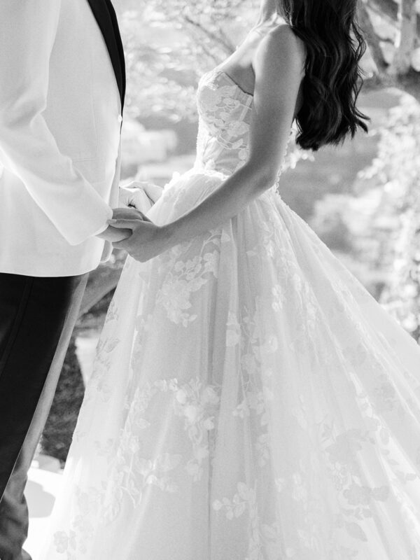 Black and white image of the couple holding hands, with the bride’s flowing gown in focus.