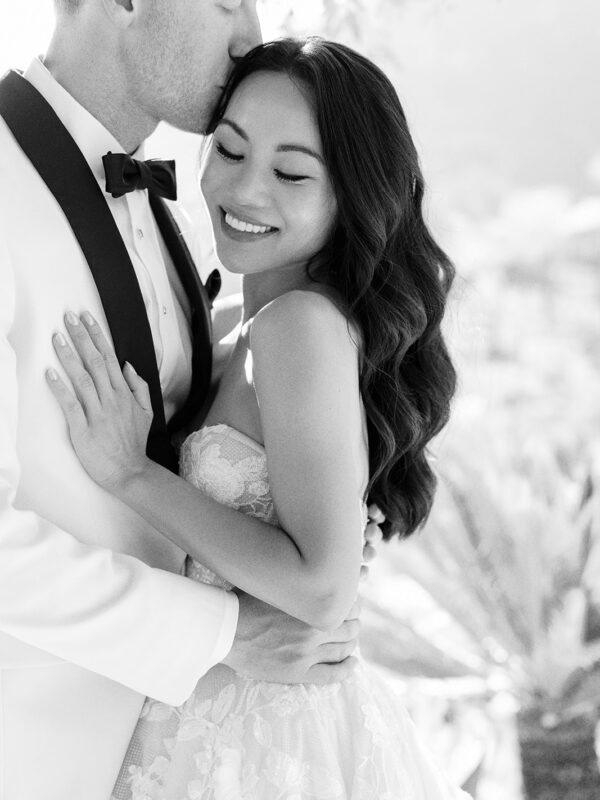 Black and white image of the groom kissing the bride’s forehead as she smiles.