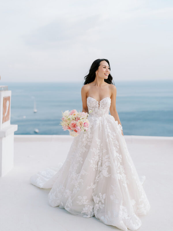 Bride smiling with the sea and sky in the background, holding a pastel bouquet.